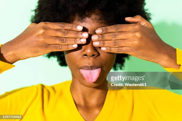 woman sticking out tongue while covering eyes with hand standing against green background - sticking out tongue stock pictures, royalty-free photos & images