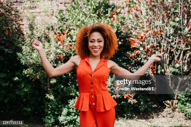 happy fashionable woman arms outstretched holding bag in public park on sunny day - jumpsuit stock pictures, royalty-free photos & images