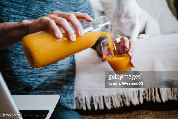 man pouring juice in glass while sitting with laptop at home - orange juice stock-fotos und bilder