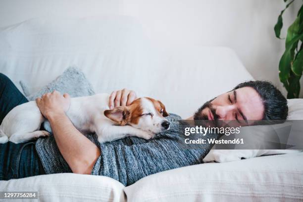 man napping with dog while lying on sofa at home - couch hund stock-fotos und bilder