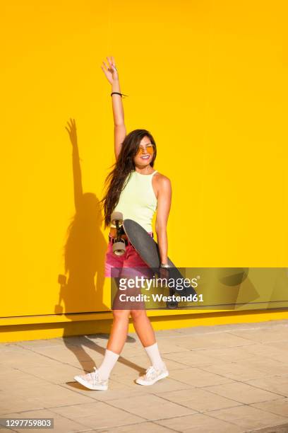 portrait of young woman walking along yellow wall with longboard in hand - pantalón corto fotografías e imágenes de stock