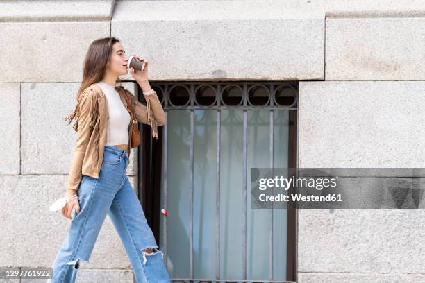 young woman drinking coffee while walking by wall - zerrissene jeans stock-fotos und bilder