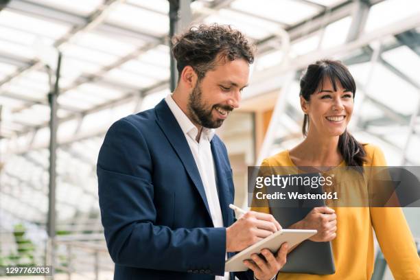 smiling male colleague using digital tablet standing by female entrepreneur at office - tablet office stock-fotos und bilder