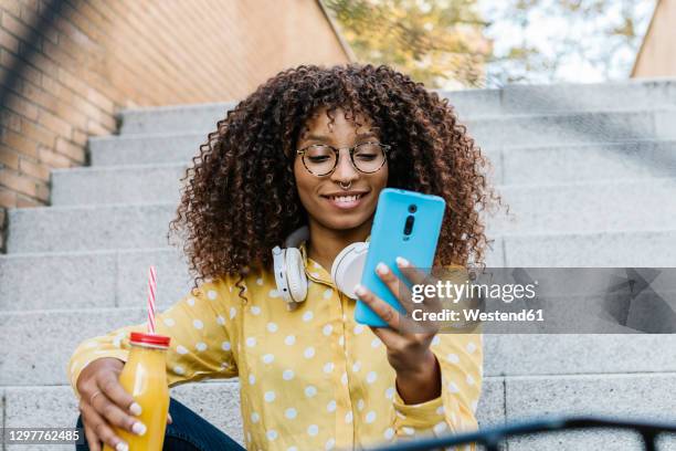 smiling woman with headphones and drink using mobile phone while sitting on steps - yellow smoothie stock pictures, royalty-free photos & images