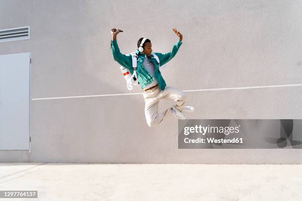 carefree man wearing headphones dancing against gray wall - in de lucht zwevend man stockfoto's en -beelden