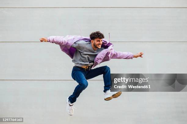 carefree young man with arms outstretched jumping against gray wall - chaqueta púrpura fotografías e imágenes de stock