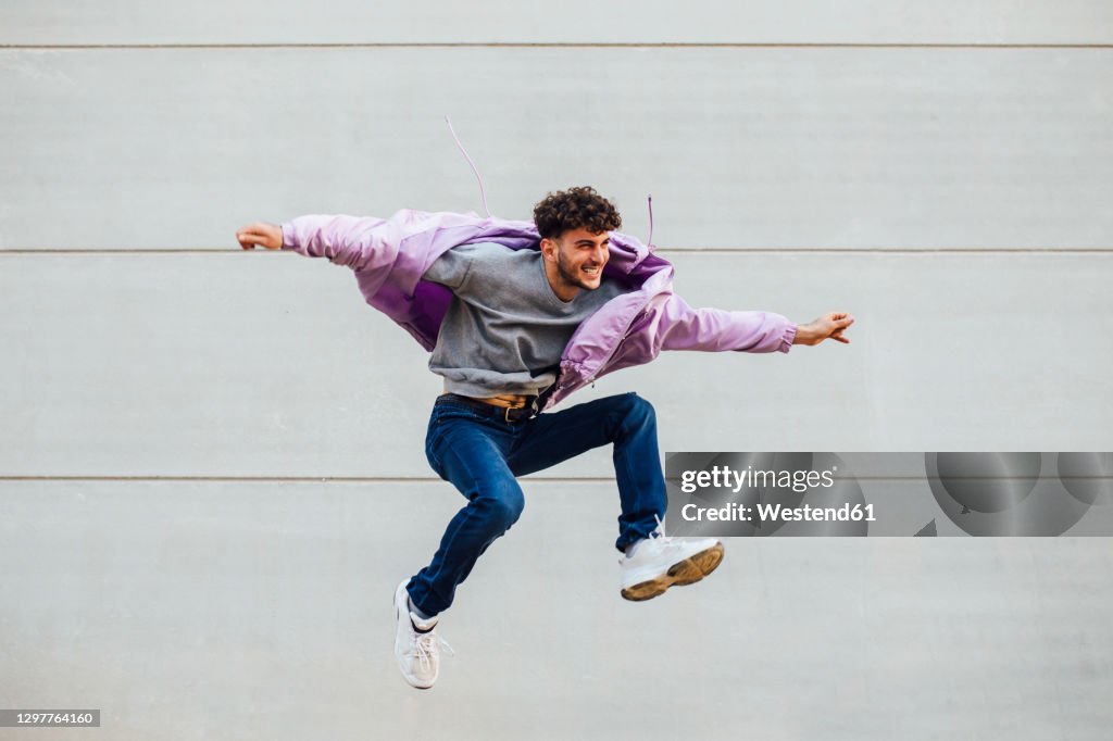 Carefree young man with arms outstretched jumping against gray wall