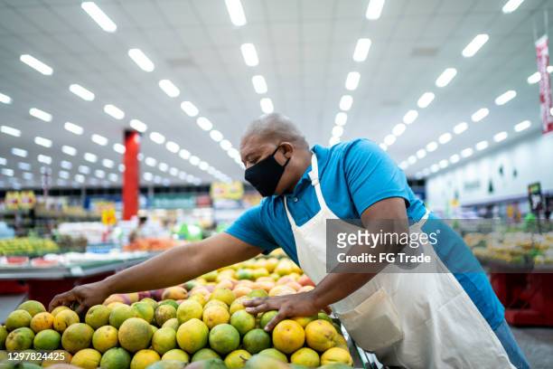 mann trägt gesichtsmaske arbeiten arrangieren obst in einem supermarkt - essenzielle berufe und dienstleistungen stock-fotos und bilder