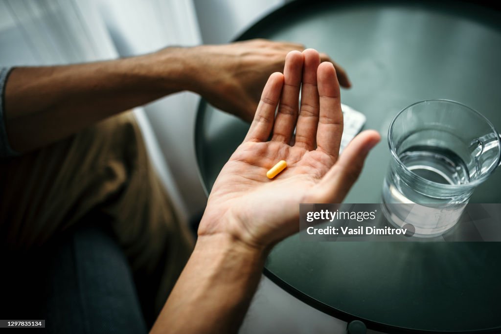 Unrecognizable man holding a pill in front of a table with glass of water. Medical treatment / drug use concept.