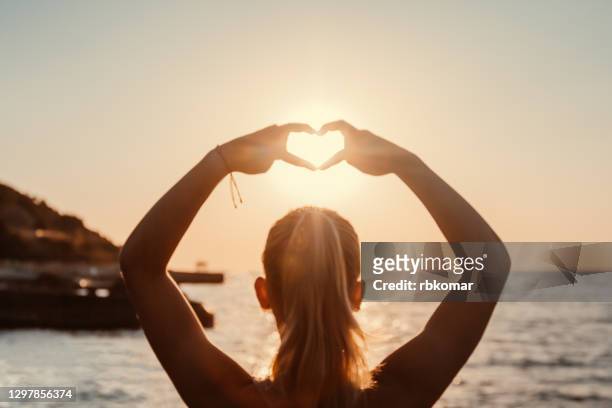 a young girl joined her raised hands in the shape of a heart against the background of the sun. female silhouette on the sea shore against the clear blue sky - aura photos et images de collection
