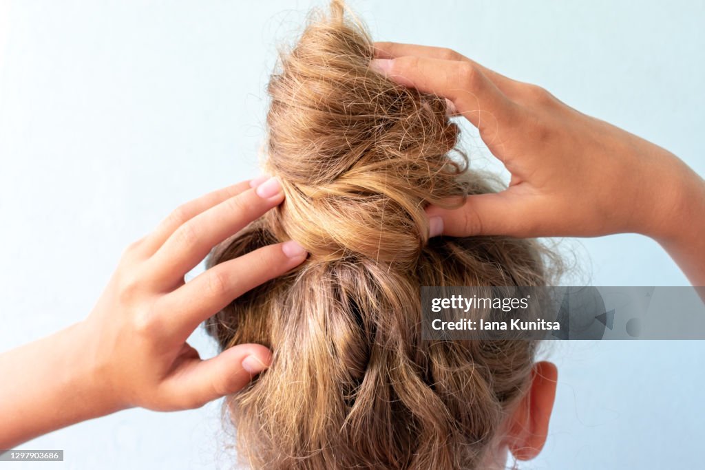 Das Mädchen richtet das entstellte Brötchen auf ihrem Kopf mit ihren Händen. Moderne schnelle Frisur. Blauer Hintergrund. Blonde lockige Haare.