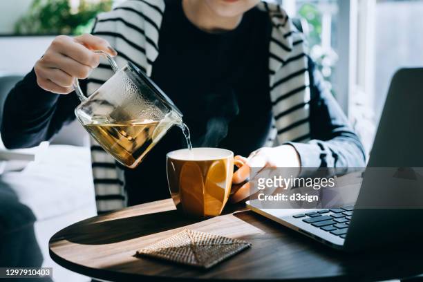 close up of young asian woman pouring a cup of tea from a stylish transparent tea pot into a cup. starting a great day ahead with a cup of hot tea while working at home on laptop in the fresh morning against sunlight. healthy lifestyle concept - fülle stock-fotos und bilder
