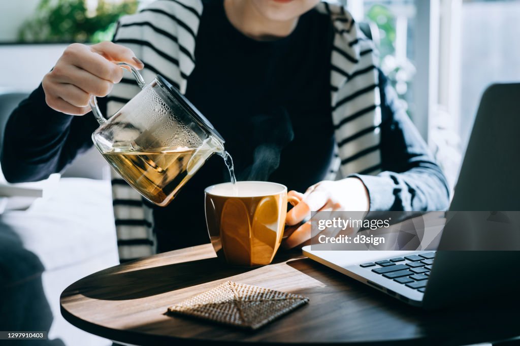 Close up of young Asian woman pouring a cup of tea from a stylish transparent tea pot into a cup. Starting a great day ahead with a cup of hot tea while working at home on laptop in the fresh morning against sunlight. Healthy lifestyle concept