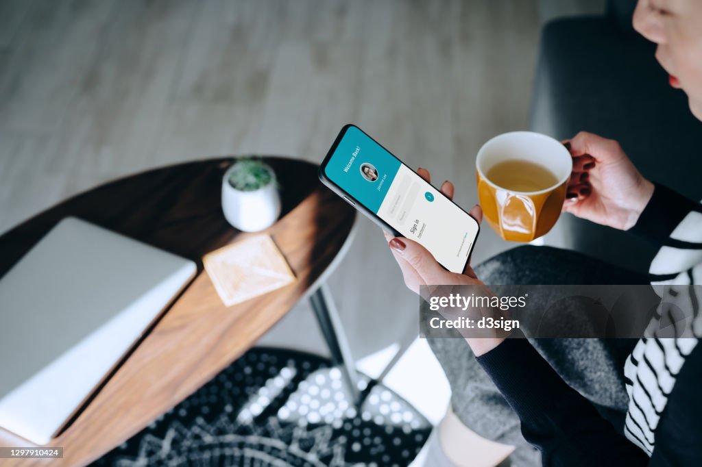 Young Asian woman relaxing at home drinking a cup of tea, logging in to her smartphone device. Lifestyle business, privacy protection, internet and mobile security concept