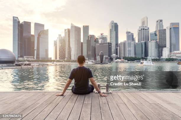 the young man sat on the dock with urban skyline and skyscrapers in marina bay singapore. - singapore imagens e fotografias de stock