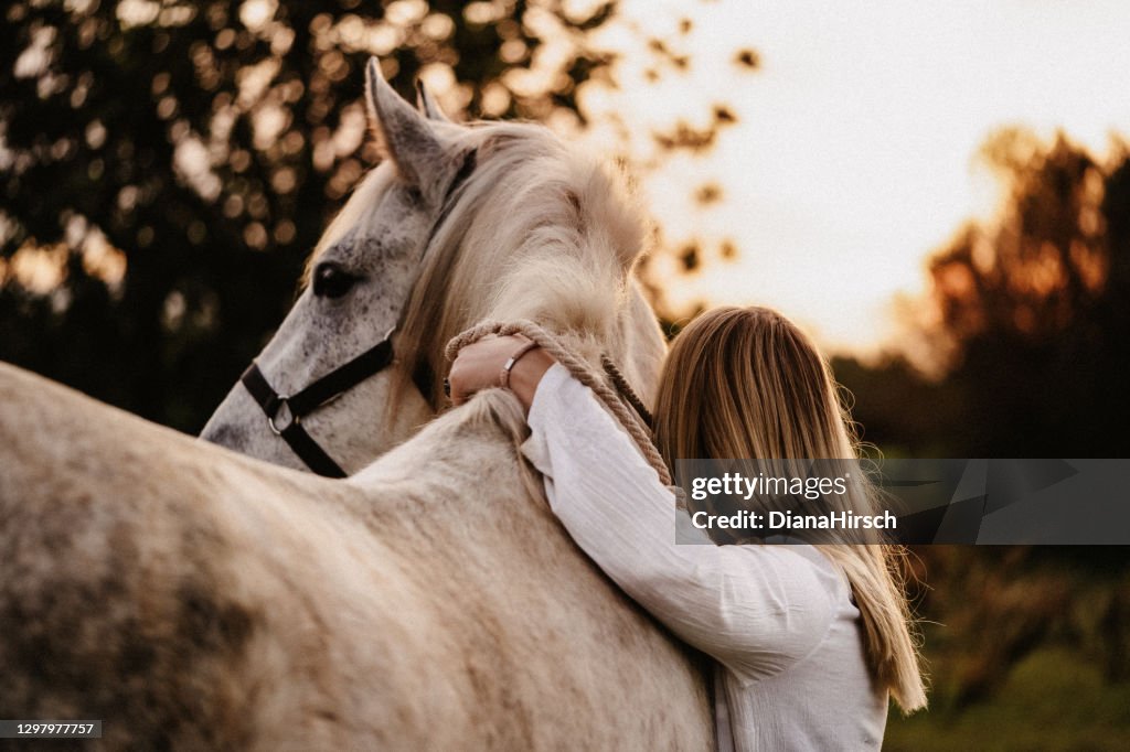 Portrait of a blond young woman from behind embracing her white horse in nature