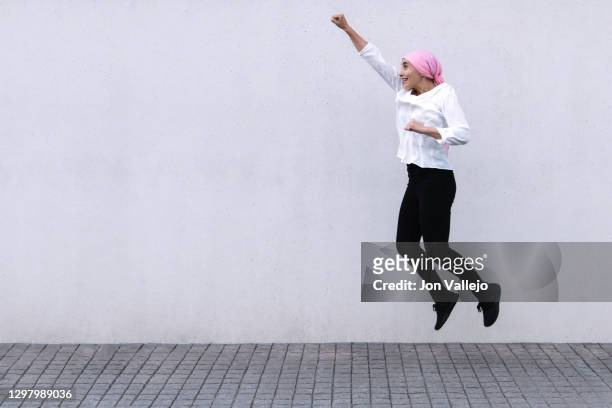 woman on the right of the image jumping with her right arm stretched and her fist closed, she is wearing a pink scarf in reference to cancer, she is wearing black pants and a white blouse. - best bosom fotografías e imágenes de stock