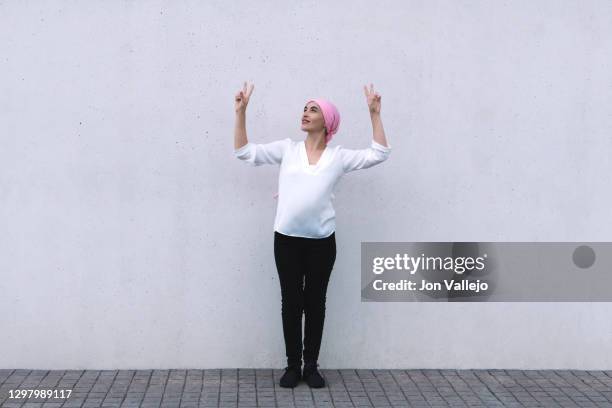 woman in the centre of the image looking up to the right, she is with both arms up making the victory sign with both hands and is wearing a pink scarf in reference to cancer, she is wearing black pants and a white blouse. - best bosom fotografías e imágenes de stock