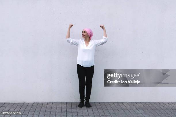 woman in the centre of the image, she is with both arms up and her fists closed as a sign of struggle and she is wearing a pink scarf in reference to cancer, she is wearing black pants and a white blouse. - cancer survivor fotografías e imágenes de stock