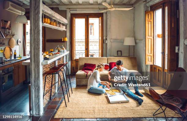 young couple sitting on floor of living room and eating pizza from box - pizza delivery stock-fotos und bilder