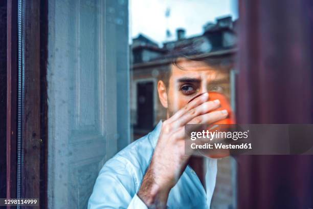 young man holding cup looking out of window - platman stockfoto's en -beelden