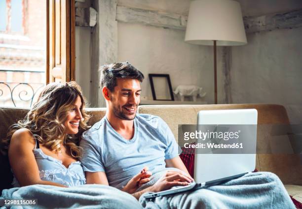 happy young couple sitting on couch at home watching laptop - lovers 2020 film fotografías e imágenes de stock