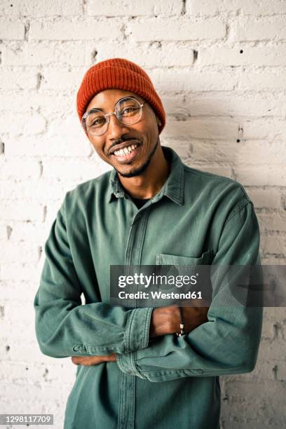 smiling man with arms crossed against white brick wall - beanie stock-fotos und bilder