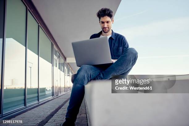 businessman using laptop while sitting on retaining wall - laptop stock-fotos und bilder