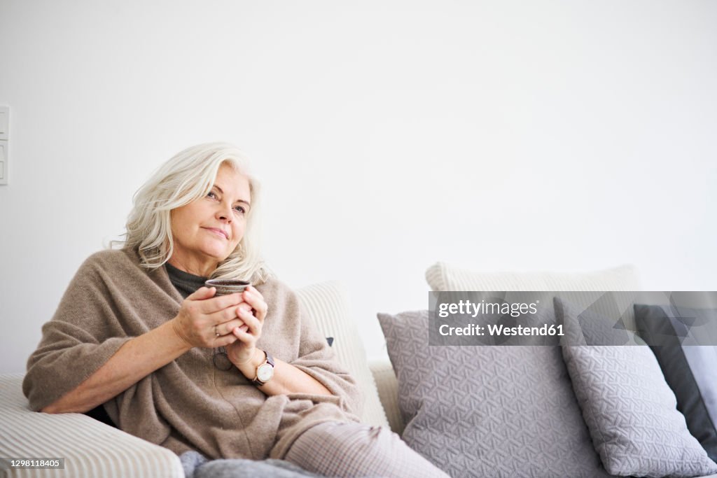 Thoughtful retired woman holding coffee cup while sitting on sofa against white wall at apartment