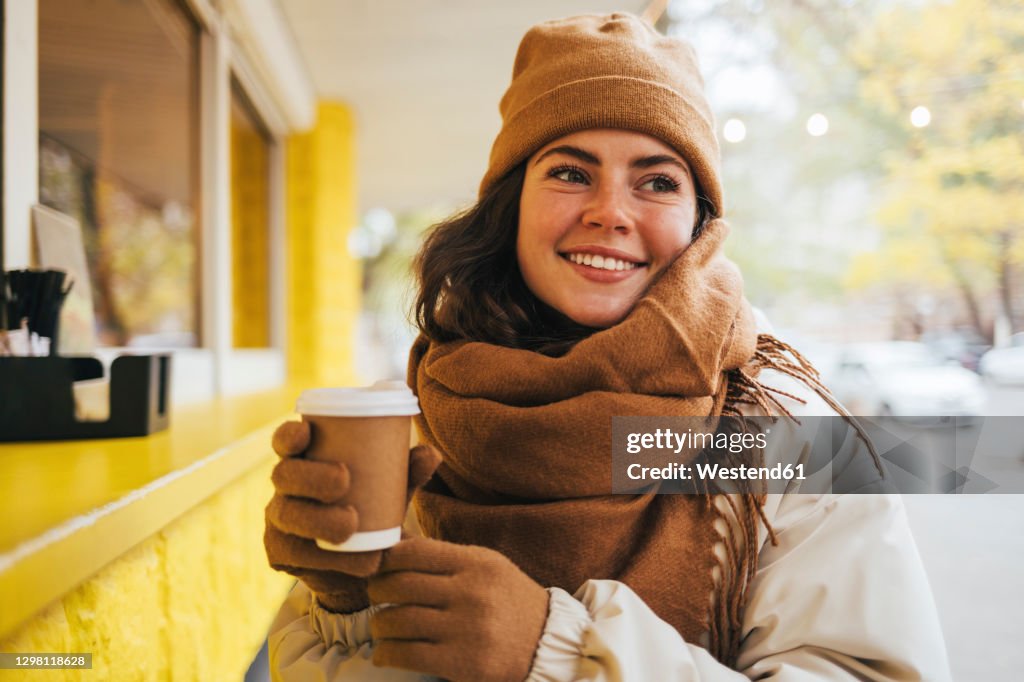 Smiling young woman looking away with disposable coffee cup at street cafe during winter