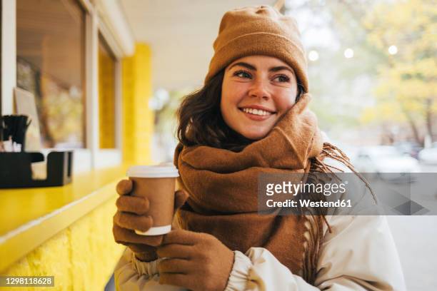 smiling young woman looking away with disposable coffee cup at street cafe during winter - woolly hat - fotografias e filmes do acervo