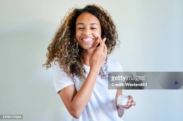 cheerful young woman applying facial cream against white wall - routine foto e immagini stock