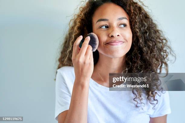 smiling young woman applying face powder with make-up brush against white wall - applying make up stockfoto's en -beelden