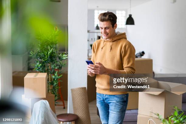 smiling young man using mobile phone in messy living room during relocation - bel appartement stockfoto's en -beelden