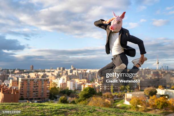 businessman wearing pig mask jumping on hill in city against sky - animal costume stock-fotos und bilder
