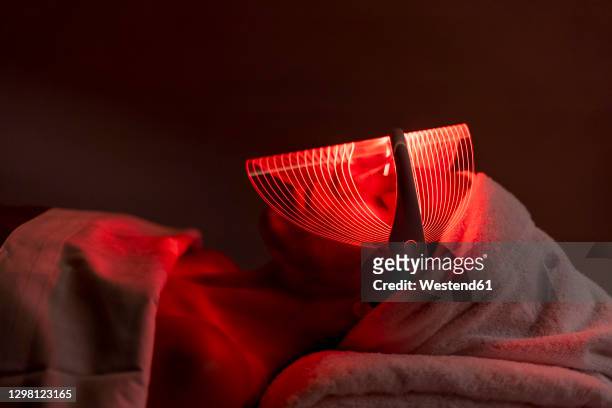 young woman wearing red mask during rejuvenating treatment in dark at beauty spa - luminothérapie photos et images de collection