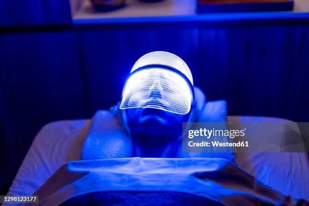 young female customer wearing illuminated blue mask during rejuvenating treatment in dark at beauty spa - blue face mask stock pictures, royalty-free photos & images