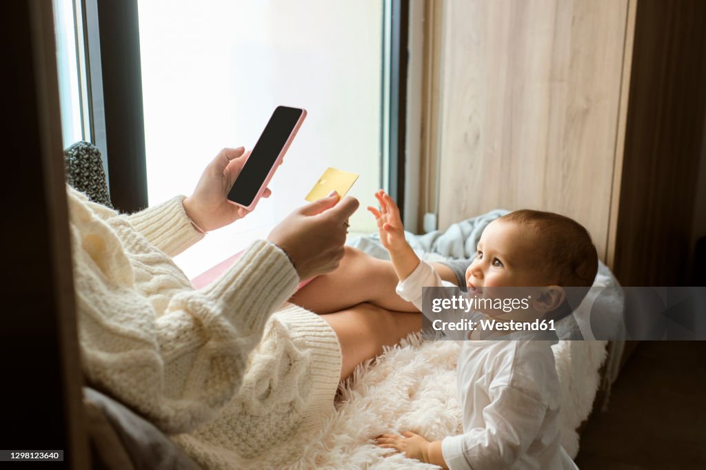 Cute baby girl reaching to credit card being held by mother sitting by window at home