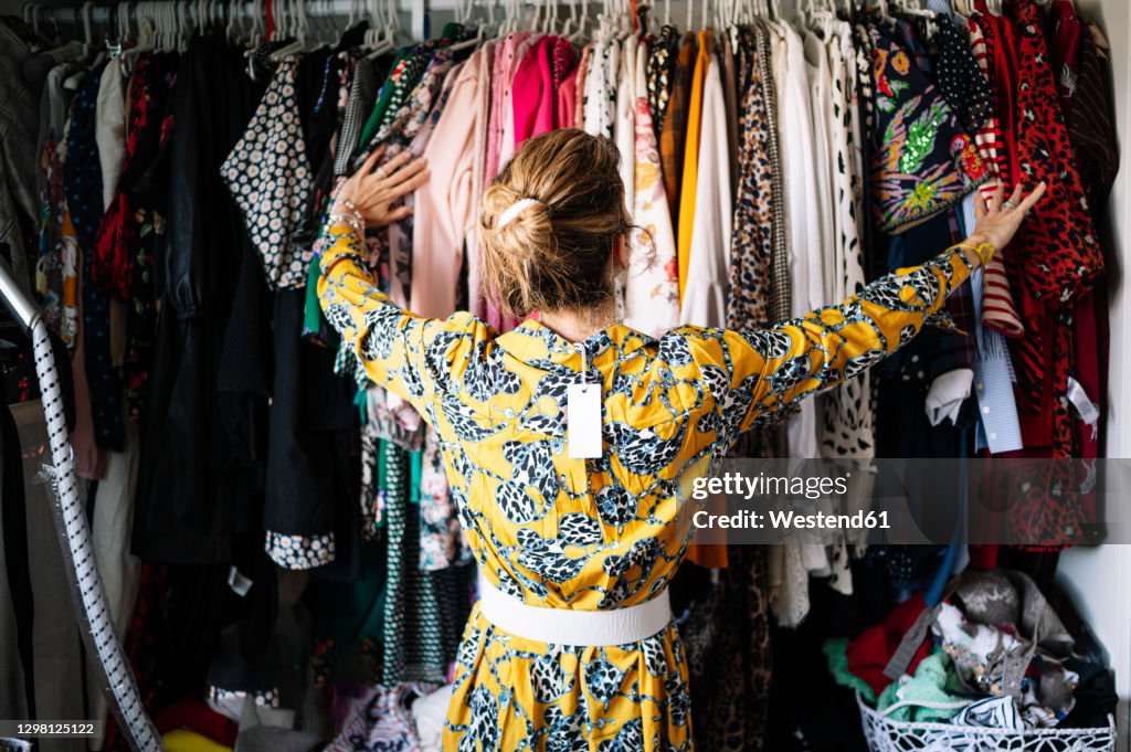 Fashionable blond woman in new yellow dress choosing from clothes rack at apartment