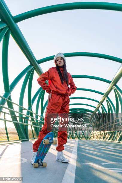 young woman with hands in pockets standing with skateboard on bridge - fashionable teen stock pictures, royalty-free photos & images