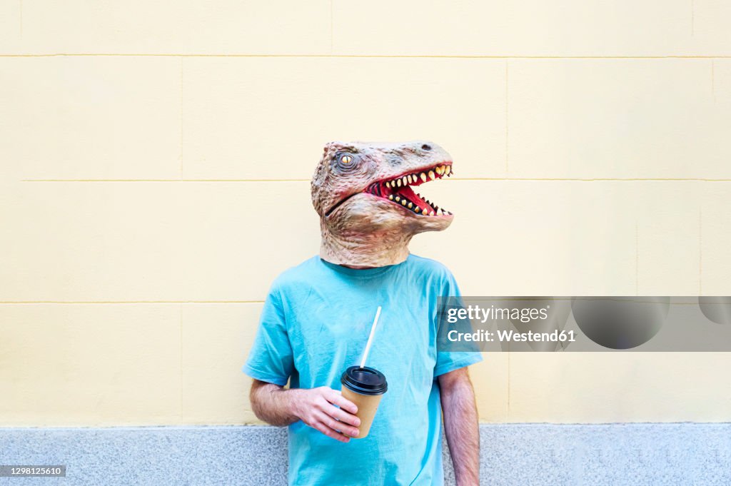 Mid adult man wearing dinosaur mask with coffee standing against wall in city