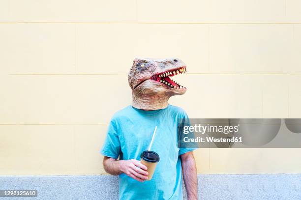 mid adult man wearing dinosaur mask with coffee standing against wall in city - man in costume stock pictures, royalty-free photos & images