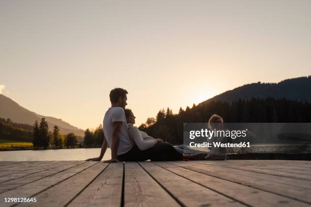 family with little daughter sitting together at end of lakeshore jetty at sunset - ponton photos et images de collection