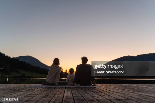 family with little daughter sitting together at end of lakeshore jetty at dusk - bergsteiger stockfoto's en -beelden