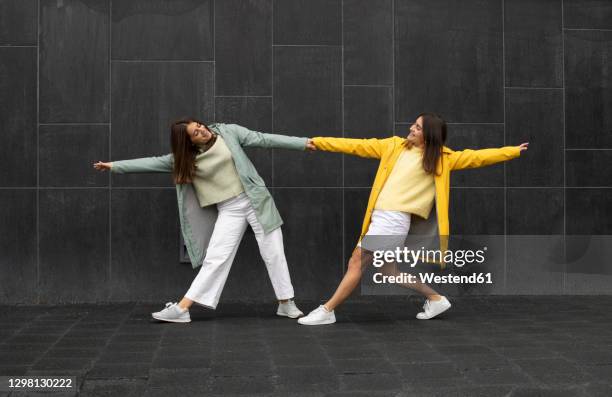 sisters wearing blue and yellow raincoats holding hands while dancing on footpath - lane sisters stockfoto's en -beelden