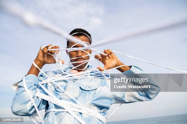 young man struggling to escape from rope while standing against sky - constraints stock-fotos und bilder