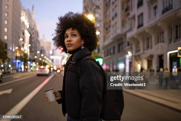 young woman with disposable coffee cup crossing road while looking around in city at night - evening walk stock-fotos und bilder