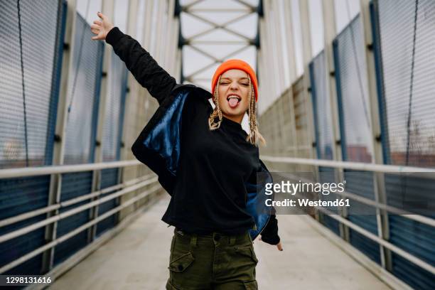 happy young woman with eyes closed sticking out tongue while dancing on footbridge - fashion stock-fotos und bilder