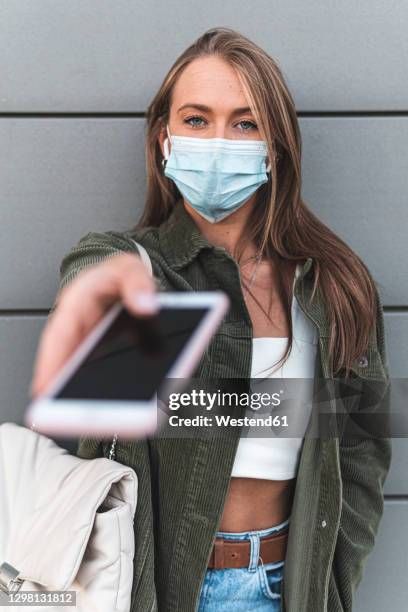 young woman wearing face mask giving smart phone while standing against wall - hand over face stock pictures, royalty-free photos & images