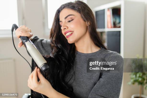 femme regardant l’ordinateur et utilisant son fer de curling tout en s’asseyant à la table d’habillage. la femme fait ses cheveux en regardant un tutoriel sur l’internet au moment de la pandémie de virus corona - fer à friser photos et images de collection